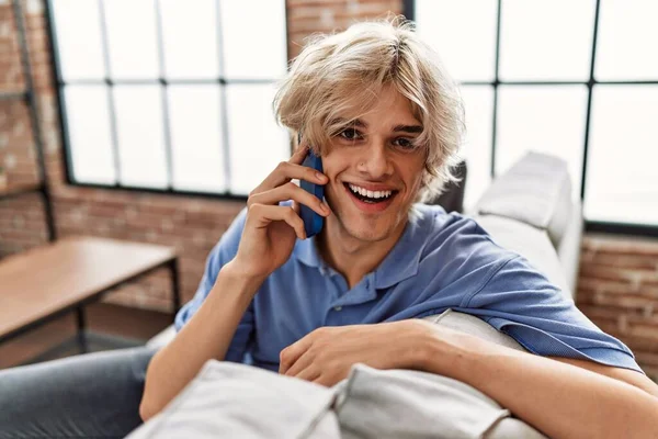 Young blond man talking on the smartphone sitting on sofa at home