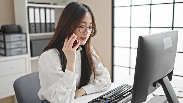 Young chinese woman business worker using computer talking on smartphone at office