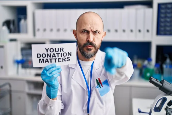 Young hispanic man working at scientist laboratory holding you donation matters banner pointing with finger to the camera and to you, confident gesture looking serious