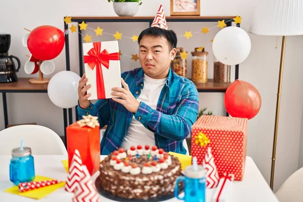Young chinese man celebrating birthday with cake and present clueless and confused expression. doubt concept.
