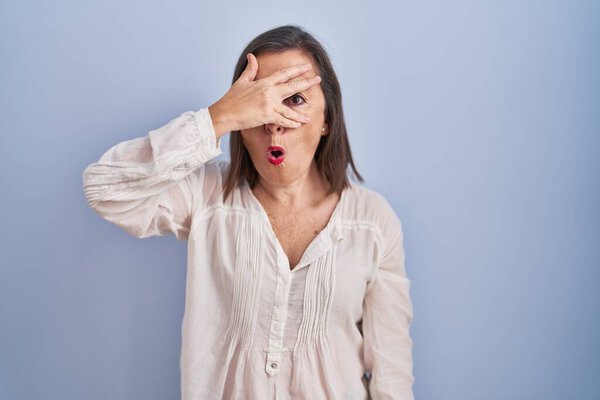 Латиноамериканка средних лет, стоящая на синем фоне, заглядывая рукой в лицо и глаза, смотрящие сквозь пальцы со смущенным выражением лица. 