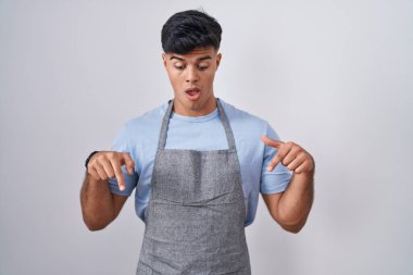 Beyaz arkaplan üzerine önlük giymiş İspanyol genç adam parmaklarını göstererek reklamını yapıyor, şaşırmış yüz ve ağzını açıyor. 