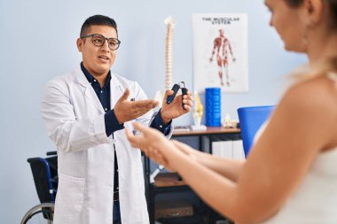 Genç latin erkek pisiyoterapist rehabilitasyon kliniğindeki hastaya el pres makinesini açıklıyor.