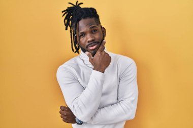 Sarı arka planda balıkçı yaka kazak giyen Afrikalı adam kameraya kollarını kavuşturmuş ve çenesini kaldırmış şekilde gülümsüyor. Olumlu düşünmek. 