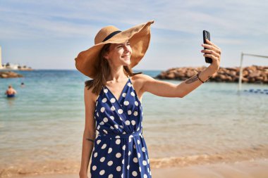 Yaz şapkası takan genç sarışın kadın sahilde akıllı telefondan selfie çekiyor.