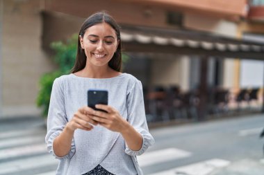 Genç, güzel, İspanyol bir kadın sokakta akıllı telefon kullanarak gülümsüyor.