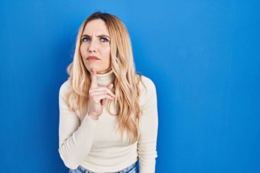 Young caucasian woman standing over blue background thinking concentrated about doubt with finger on chin and looking up wondering 