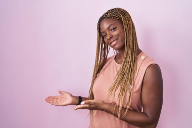 African american woman with braided hair standing over pink background inviting to enter smiling natural with open hand 