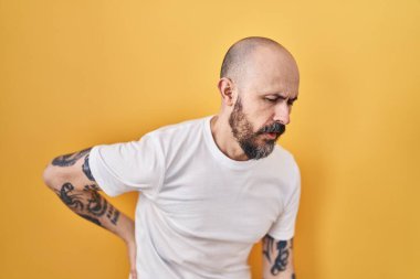 Sarı arka planda duran İspanyol dövmeli genç adam sırt ağrısı çekiyor, elleriyle dokunuyor, kas ağrısı çekiyor. 