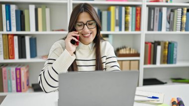 Young beautiful hispanic woman student using computer talking on smartphone at library university