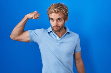 Mavi arka planda duran beyaz adam güçlü kol kası gösteren, kendinden emin ve gururlu bir adam. 