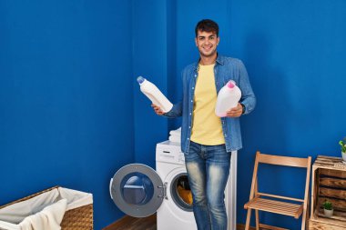 Çamaşır odasında çamaşır deterjanı şişeleriyle çamaşır yıkayan İspanyol genç adam.