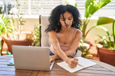 Young hispanic woman sitting on table studying at home terrace