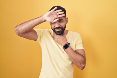 Hispanic man with beard standing over yellow background touching forehead for illness and fever, flu and cold, virus sick 
