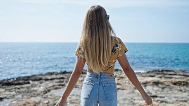 Genç sarışın kadın deniz kenarında kollarını ters açmış duruyor.