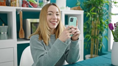 Genç sarışın kadın yemek odasındaki masada oturan akıllı telefonu kullanıyor.