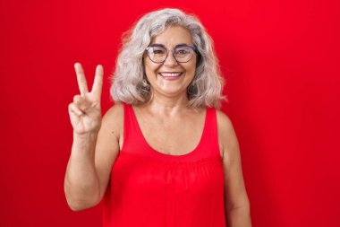 Kırmızı arka planda gri saçlı orta yaşlı bir kadın iki numaralı parmaklarını gösterip gülümserken kendinden emin ve mutlu görünüyor.. 