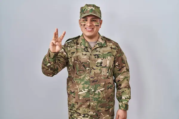 Hispanic young man wearing camouflage army uniform showing and pointing up with fingers number three while smiling confident and happy.