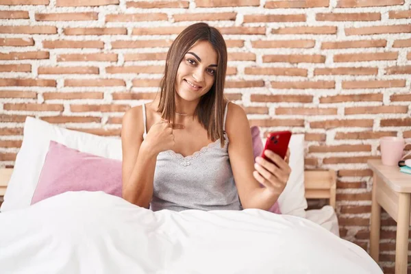 自宅でベッドに座ってスマートフォンを使用する若いヒスパニックの女性は 幸せで誇りに思える1つの自己を指差します — ストック写真