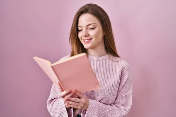年轻的高加索女人正在阅读一本粉红背景的书 脸上带着快乐而冷静的微笑 显示牙齿 — 图库照片