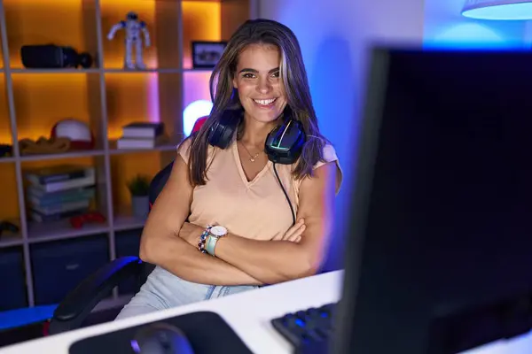 Young beautiful hispanic woman streamer smiling confident sitting with arms crossed gesture at gaming room