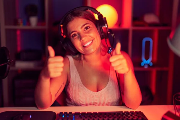 Young blonde woman playing video games wearing headphones success sign doing positive gesture with hand, thumbs up smiling and happy. cheerful expression and winner gesture.