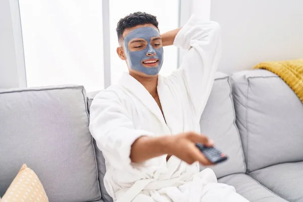 Young latin man relaxed on sofa with facial mask treatment watching tv at home