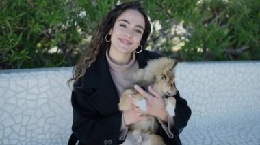 Köpeğiyle gülümseyen genç İspanyol kadın parkta bankta oturuyor.