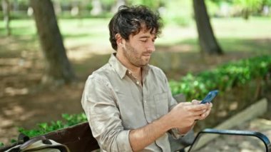 Parktaki bankta oturan akıllı telefonu kullanarak kendine güvenen İspanyol genç adam.