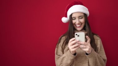 Güzel İspanyol kadın, yılbaşı şapkası takıyor ve izole edilmiş kırmızı arka plan üzerinde akıllı telefon kullanıyor.