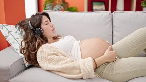 若いです妊娠中の女性聞いています音楽に寝ますオンソファにホーム — ストック動画