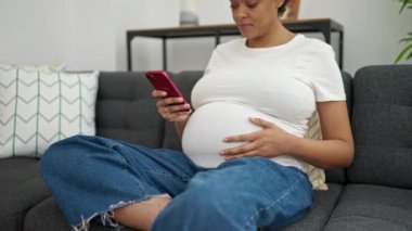 Evdeki kanepede oturan akıllı telefon kullanan hamile bir kadın.