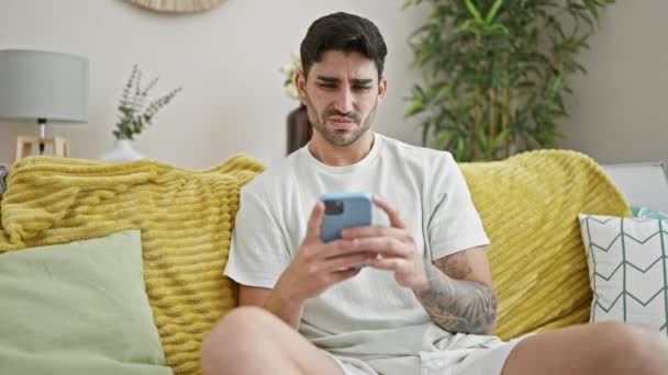 Νεαρός Ισπανόφωνος Άνδρας Χρησιμοποιώντας Smartphone Κάθεται Στον Καναπέ Αναζητούν Αναστατωμένος — Αρχείο Βίντεο