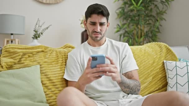 Ung Latinamerikan Man Med Smartphone Sitter Soffan Ser Upprörd Hemma — Stockvideo