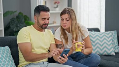 Kadın ve erkek, evdeki kanepede oturmuş akıllı telefon kullanarak kahve içiyorlar.
