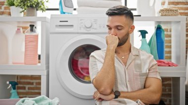 Çamaşır makinesine yaslanan genç İspanyol adam çamaşır odasında stres altında.