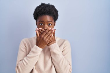 African american woman standing over blue background shocked covering mouth with hands for mistake. secret concept. 