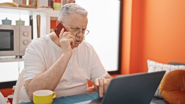 Yemek odasında bilgisayarla konuşan orta yaşlı, kır saçlı bir adam.