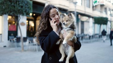 Köpeğiyle gülümseyen Latin kökenli genç bir kadın sokakta akıllı telefondan konuşuyor.