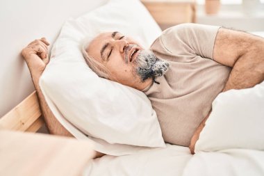Orta yaşlı, kır saçlı bir adam yatakta uzanıyor ve yatak odasında uyuyor.