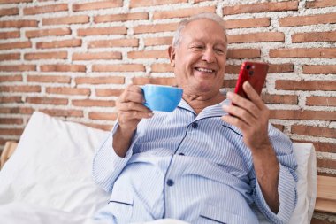 Orta yaşlı, kır saçlı, akıllı telefon kullanan, yatak odasında kahve içen bir adam.