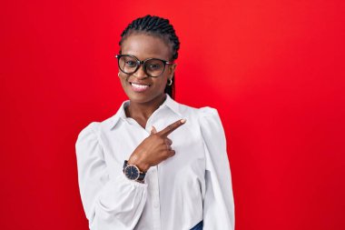 Örgü ören, kırmızı arka planda neşeli, yüzünde mutlu ve doğal bir ifadeyle elleri ve parmaklarıyla gülümseyen Afrikalı bir kadın. 