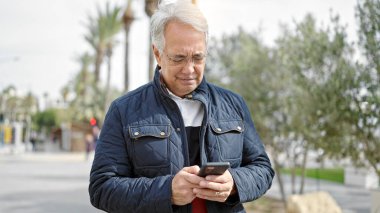 Parkta akıllı telefon kullanan gri saçlı orta yaşlı bir adam.