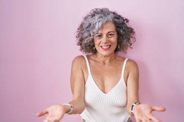 Pembe arka planda duran gri saçlı orta yaşlı kadın. Kolları açık gülümsüyor. Dostça karşılama, olumlu ve kendinden emin selamlar. 