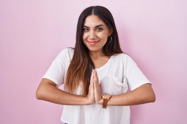 Pembe arka planda duran genç bir Arap kadın ellerini birleştirip af diliyor kendinden emin bir şekilde gülümsüyor.. 