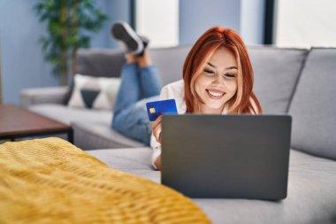 Evdeki kanepede dizüstü bilgisayar ve kredi kartı kullanan beyaz bir kadın.