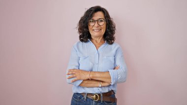 Orta yaşlı, gözlüklü, kolları çapraz pembe arka planda duran İspanyol bir kadın.