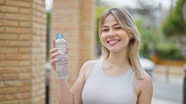 Genç sarışın kadın gülümsüyor. Elinde bir şişe su tutuyor.