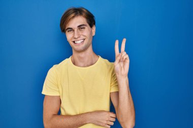 Mavi arka planda duran genç adam kameraya bakıp gülümsüyor ve zafer işareti yapıyor. İki numara.. 