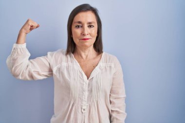 Orta yaşlı Latin bir kadın mavi arka planda duruyor güçlü bir insan kol kası gösteriyor, kendinden emin ve gururlu. 